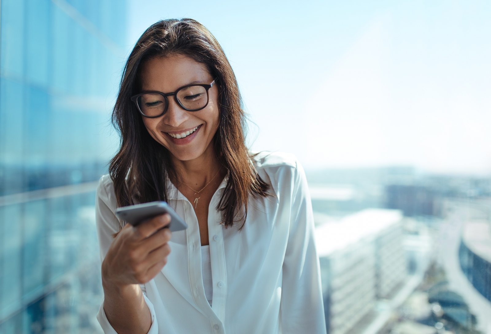 Woman wearing eyeglasses smiling while looking at her mobile phone. Young businesswoman using mobile phone for business communication.