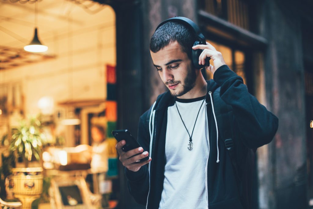 Hispanic young attractive man stands in dark street in front of shop changes songs and tracks on smartphone listens to music in wireless headphones. Hipster with slight beard