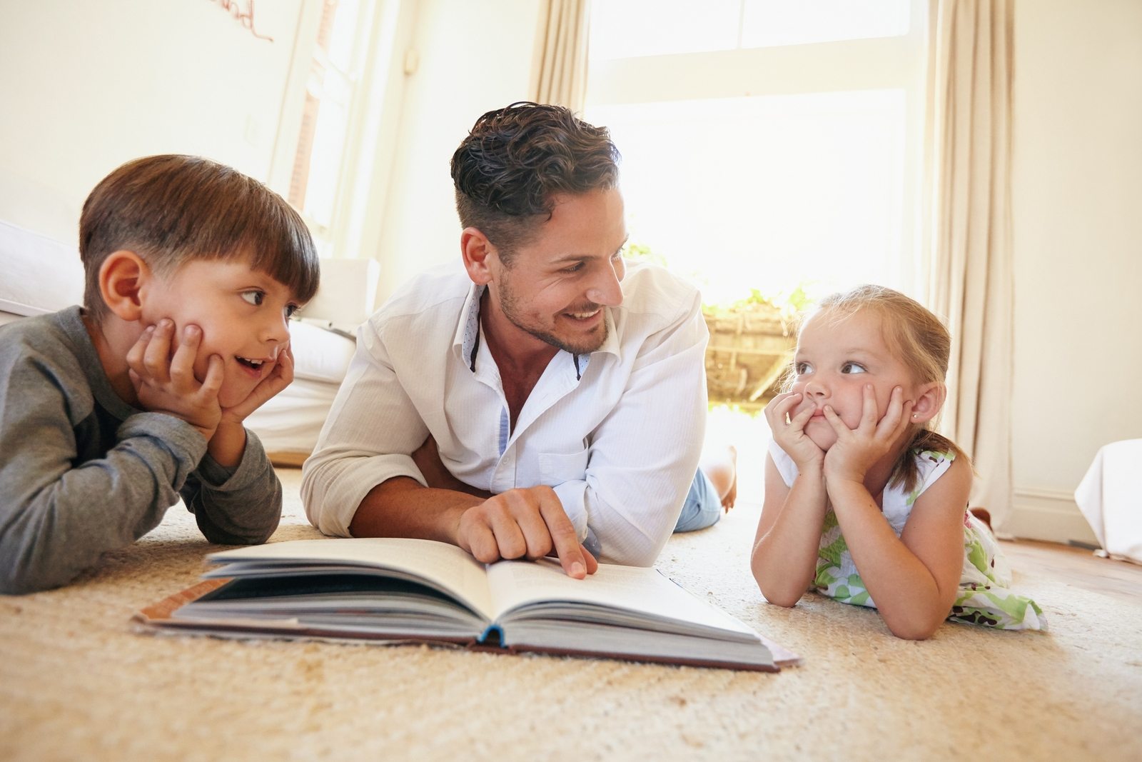 Indoor shot of young man with two kids reading a story book . Family lying on the floor with a book in living room.